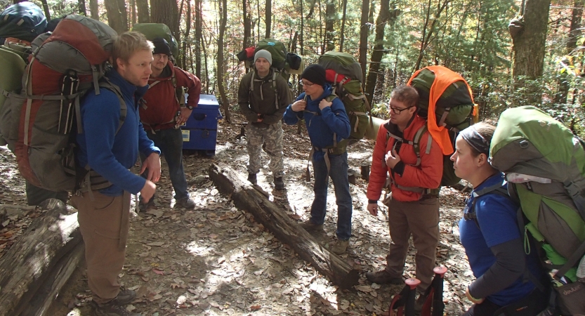 A group of backpackers talk to each other in a wooded area. 